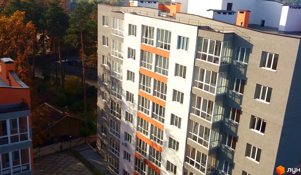 Зовнішній вигляд житлового комплексу Status з білими та сірими фасадами, помаранчевими вставками та великими вікнами. Навколо комплексу зелені дерева та частина прибудинкової території.