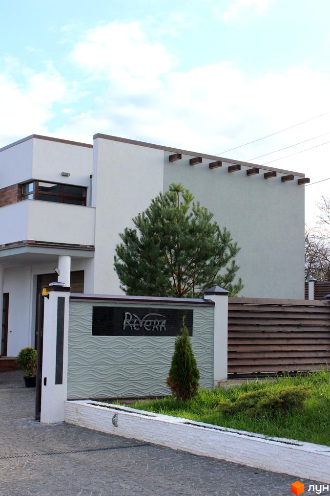 Зовнішній вигляд котеджного містечка Riviera з фасадом білого кольору та дерев'яними елементами. Прибудинкова територія з зеленими насадженнями та парканом.