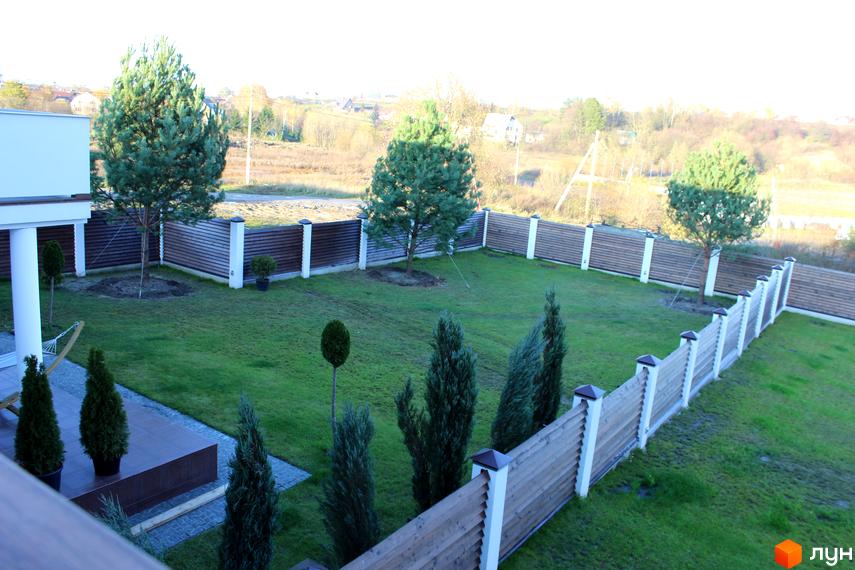 Котеджне містечко Riviera: оглядовий вид на зелену галявину з деревами та парканом. На території житлового комплексу облаштована прибудинкова територія з газоном та декоративними рослинами.