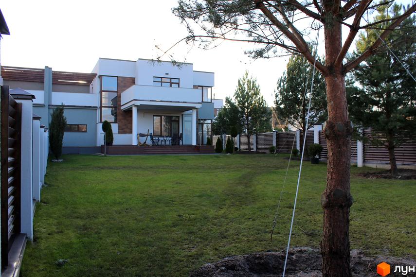 Котеджне містечко КМ Riviera. Зовнішній вигляд двоповерхового будинку з великими вікнами та терасою, зелена галявина та дерева на прибудинковій території.