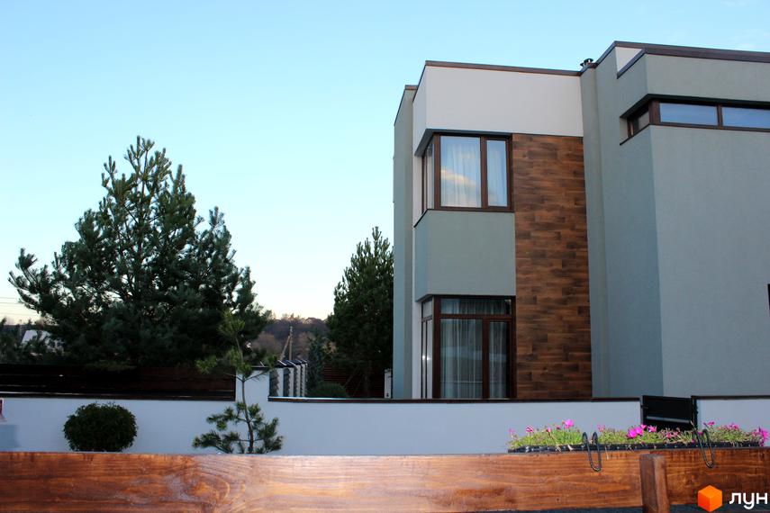 Котеджне містечко Riviera: двоповерховий будинок з коричневими та сірими фасадами, великими вікнами та зеленою прибудинковою територією з деревами та квітами.