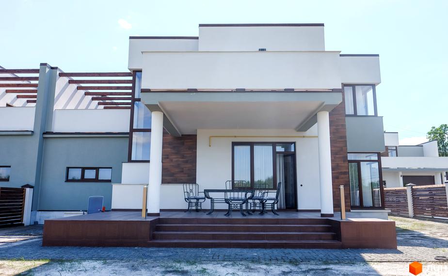 Котеджне містечко Riviera. Зовнішній вигляд двоповерхового будинку з терасою, колонами та дерев'яною обшивкою. На терасі стоять столики та стільці.