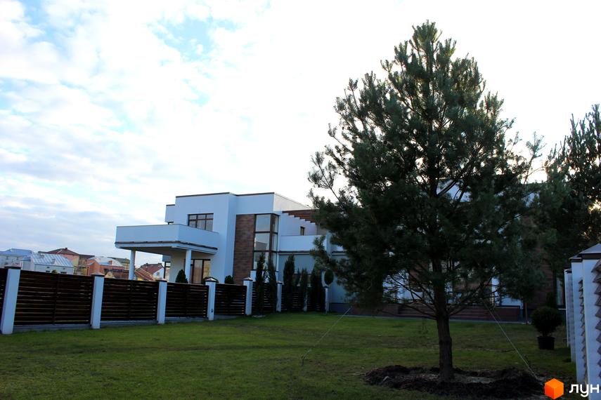 Котеджне містечко КМ Riviera: білосніжний двоповерховий будинок з великими вікнами та балконом, огороджена територія з доглянутим газоном та деревами.