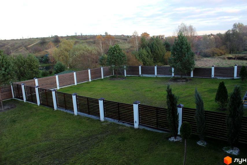 Котеджне містечко Riviera з дерев'яним парканом та зеленим газоном. На задньому плані пагорби з будинками та деревами.