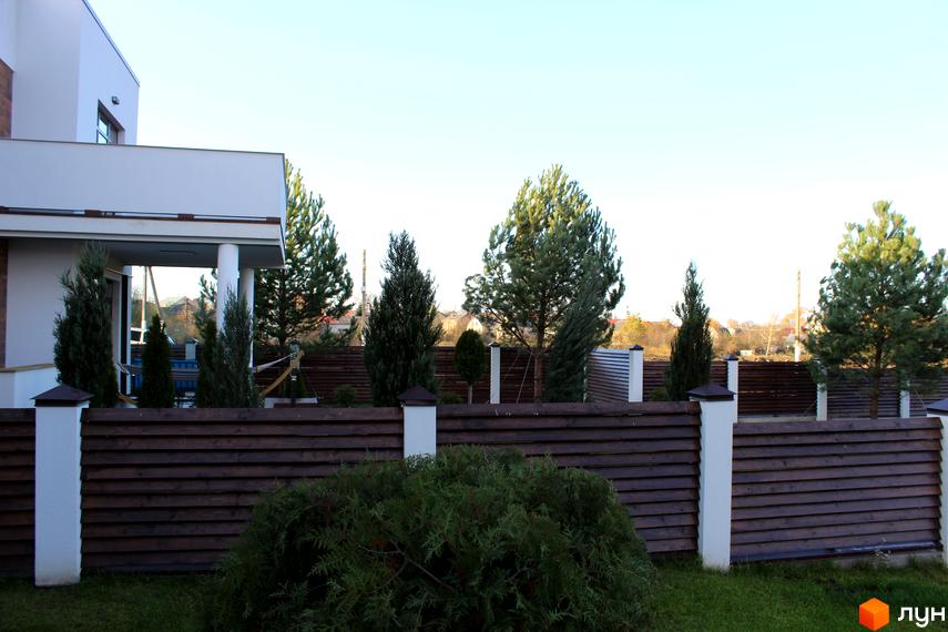 Котеджне містечко Riviera: сучасний фасад будинку з колонами та дерев'яною огорожею. Прибудинкова територія з туями та соснами.