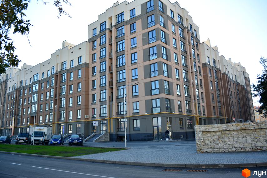 Багатоповерховий житловий комплекс Барселона з бежево-сірим фасадом та численними вікнами. Перед ним вулиця з автомобілями та зеленою зоною.