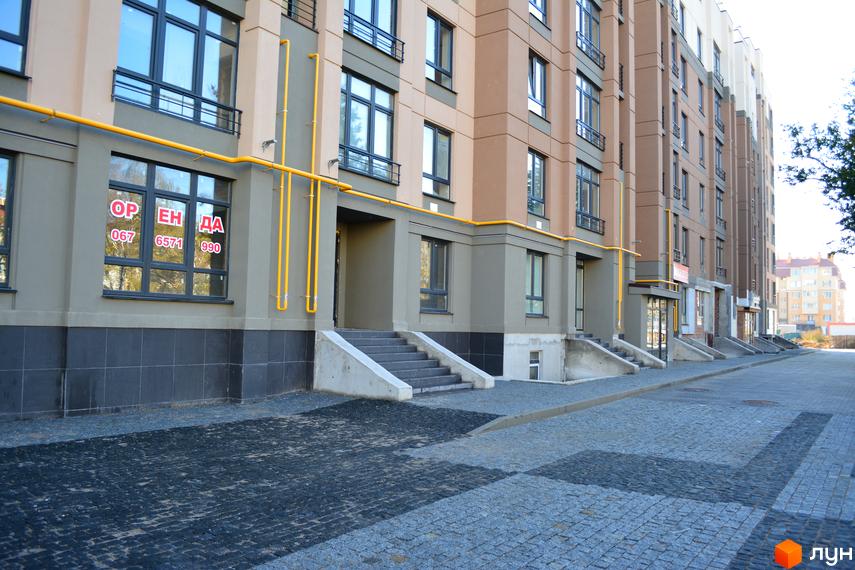 Зовнішній вигляд новобудови ЖК Барселона з сірими фасадами та жовтими комунікаціями. Прибудинкова територія з бруківкою та сходами до під'їздів.