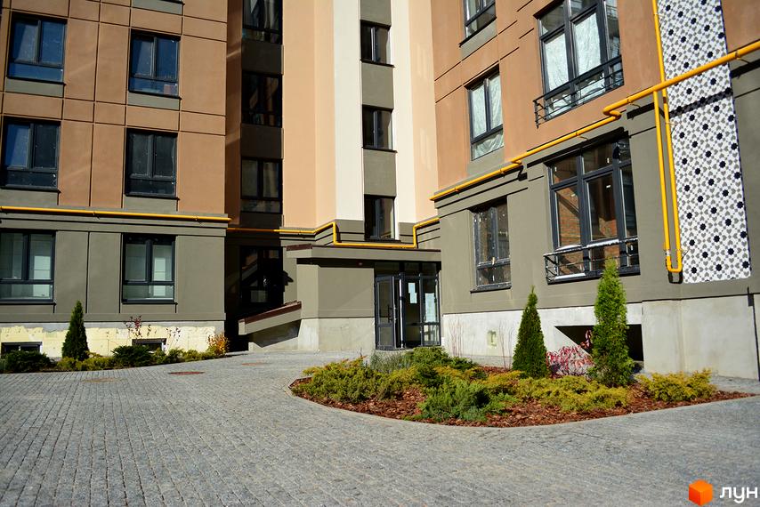 Зовнішній вигляд будинку ЖК Барселона з сірим мощенням та зеленими насадженнями. Сучасна архітектура та доглянута прибудинкова територія.