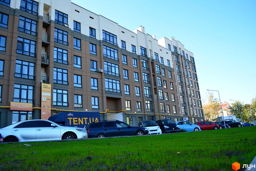 Багатоповерховий будинок ЖК Барселона з коричнево-сірими фасадами та вікнами. Перед ним припарковані авто на тлі зеленої зони.