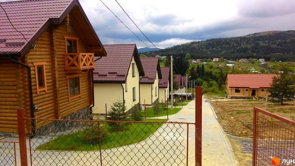 Новобудова на вулиці Грушевського, сучасні дерев'яні будинки з червоними дахами та доглянутою прибудинковою територією. Зелені пагорби та хмарне небо на фоні.