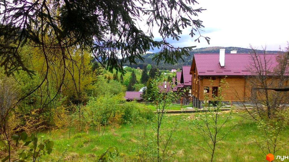 Будинок з дерев'яних колод під червоним дахом у зеленій місцевості. Навколо будинку розташовані дерева та трава, що створює затишну атмосферу.