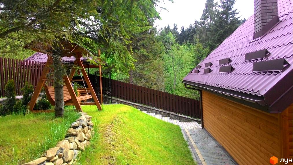 Зовнішній вигляд будинку з дерев'яними стінами та фіолетовим дахом. Прибудинкова територія з гойдалкою та кам'яною огорожею.