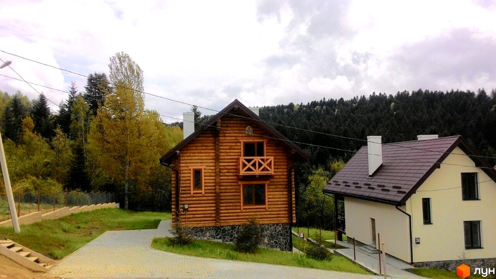 Двоповерховий дерев'яний будинок з балконом та сусідній світлий будинок під коричневим дахом. Навколо будинків зелена територія та дерева.