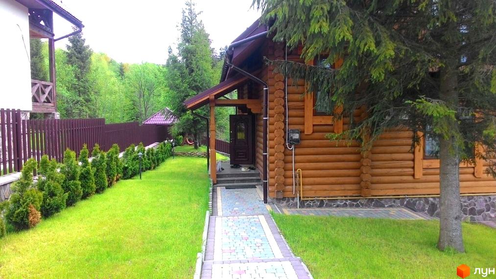 Затишний дерев'яний будинок з терасою та доглянутою прибудинковою територією, вимощеною доріжкою та зеленим газоном. Навколо будинку розташовані дерева та огорожа.