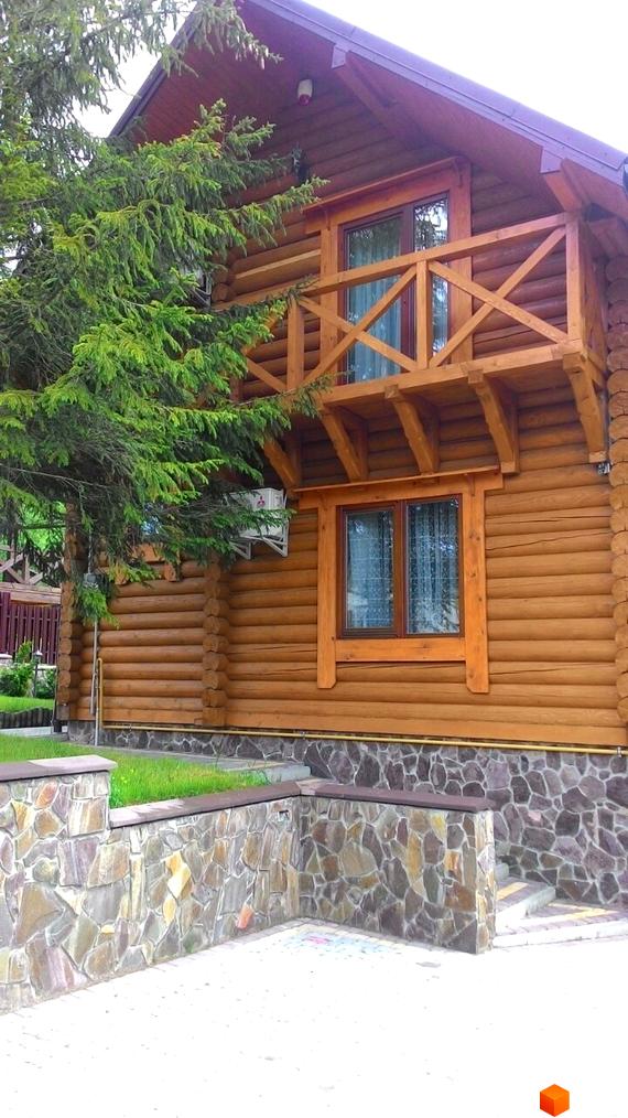 Дерев'яний будинок з балконом та вікнами, оточений зеленню. Навколо будинку кам'яна огорожа та бруківка.