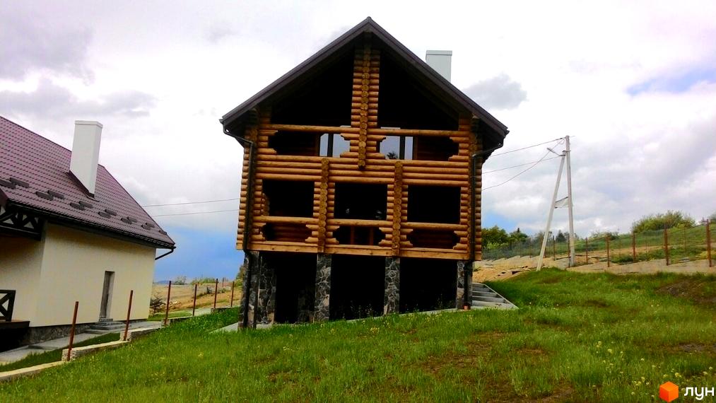 Двоповерховий дерев'яний будинок з великими вікнами та кам'яним цоколем. Навколо будинку зелена трава та паркан.