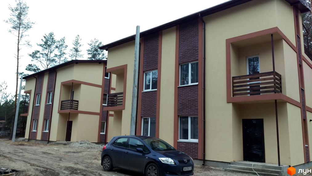 Зображення житлового комплексу. На фото будівля з бежевими стінами та коричневими вставками з цегли. Перед будинком припаркований автомобіль, навколо територія з гравієм.