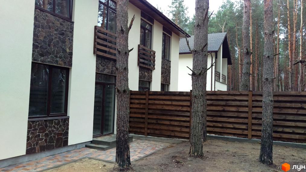 Котеджне містечко КМ Лісове-3: двоповерховий будинок з кам'яним оздобленням фасаду та дерев'яними балконними огородженнями. Прибудинкова територія з дерев'яним парканом та соснами.