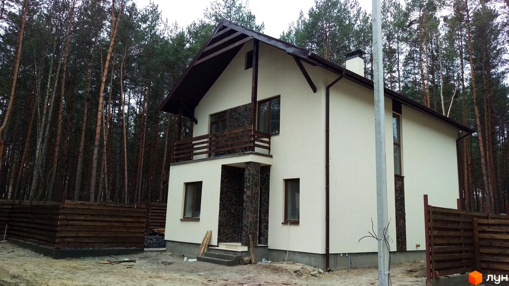 Котеджне містечко КМ Лісове-3. Зовнішній вигляд двоповерхового будинку з коричневим дахом та дерев'яним балконом на тлі соснового лісу. Прибудинкова територія огороджена дерев'яним парканом.