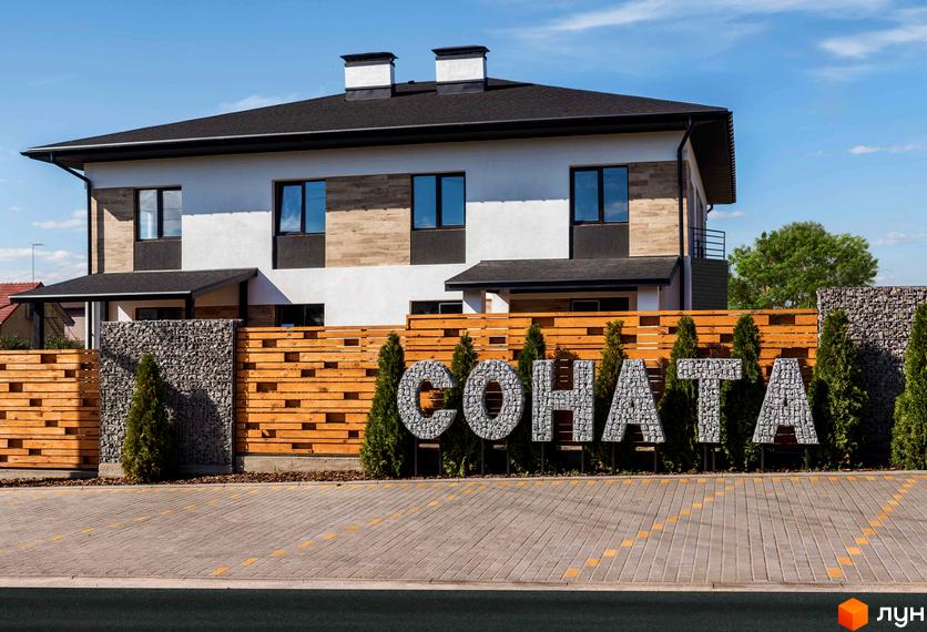 Котеджне містечко Соната. Фасад будинку з білою та дерев'яною обробкою, великими вікнами. Прибудинкова територія з дерев'яним парканом, кам'яними літерами та туями.