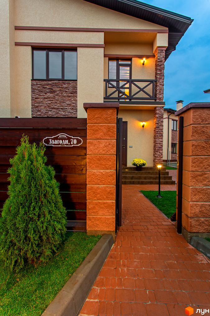 Котеджне містечко Злагода: будинок з бежевим фасадом, кам'яним декором та дерев'яним парканом. Вхідна група з бруківкою та зеленим газоном.