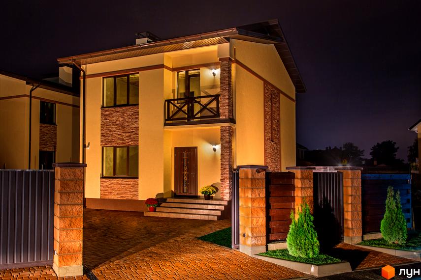Котеджне містечко КМ Злагода. Двоповерховий будинок з жовтим фасадом, кам'яним оздобленням та балконом, освітлений вночі. Прибудинкова територія з бруківкою та зеленими насадженнями.