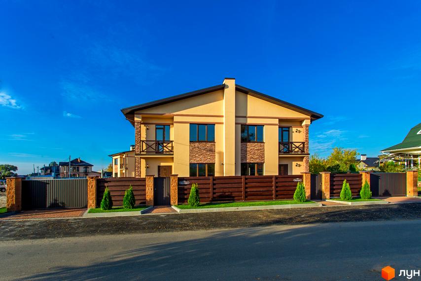 Котеджне містечко Злагода: двоповерховий будинок світло-бежевого кольору з коричневим дахом та кам'яним оздобленням фасаду. Огороджена територія з парканом та зеленими насадженнями.