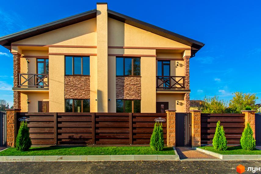 Котеджне містечко Злагода: двоповерховий будинок світло-жовтого кольору з коричневими вікнами та балконами, огороджений дерев'яним парканом. Прибудинкова територія доглянута, з газоном та туями.