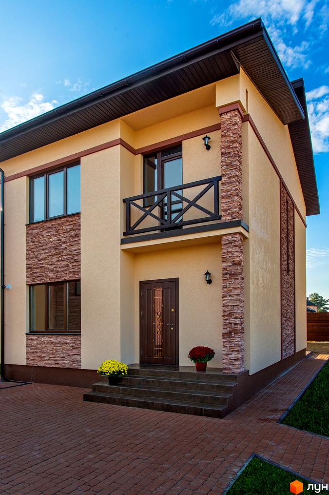 Котеджне містечко КМ Zlagoda. Фасад двоповерхового будинку з бежевою штукатуркою, коричневими вікнами та дверима, декоративними кам'яними колонами. Біля будинку вимощена доріжка та зелена галявина.