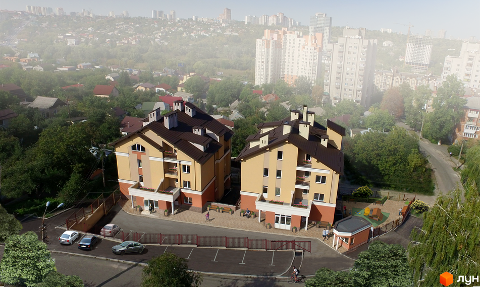 Житловий комплекс Comfort Club - двоповерхові будинки світло-жовтого кольору з коричневими дахами. Поруч дитячий майданчик та парковка з автомобілями.