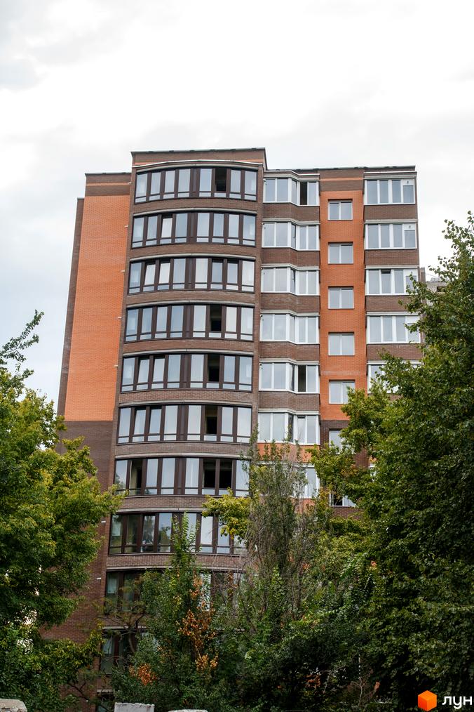 Зовнішній вигляд житлового комплексу Ковалівський з цегляним фасадом та великими вікнами. Навколо будинку зелені дерева та рослинність.