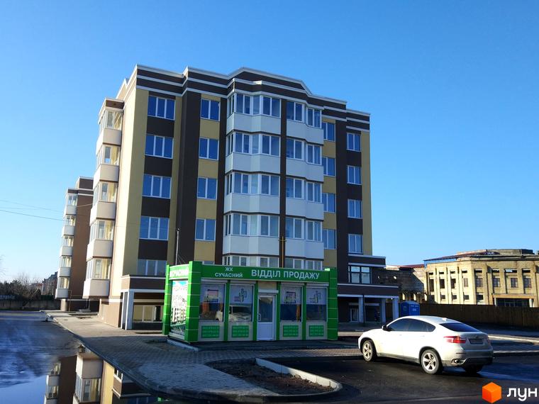 Житловий комплекс Сучасний - це багатоповерховий будинок з балконами та великими вікнами. Перед будинком розташована прибудинкова територія з дорогою та автомобілем.