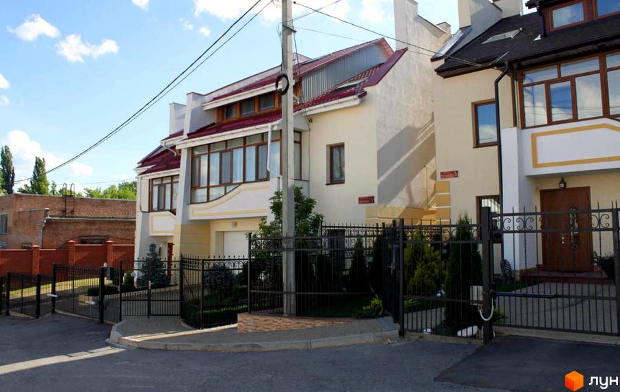 Зображення двоповерхового будинку з червоним дахом та великими вікнами. Перед будинком металева огорожа та зелені насадження.