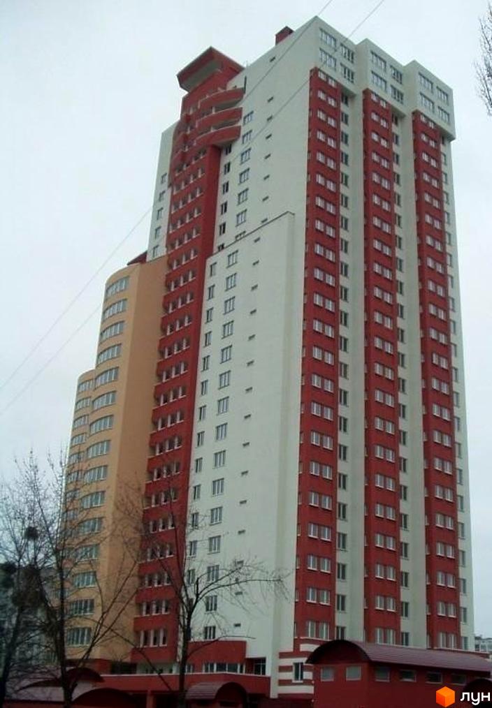 Багатоповерховий житловий комплекс на вул. Тичини, 18б, з біло-червоним фасадом та прилеглою бежевою секцією. На передньому плані видно дерева без листя.