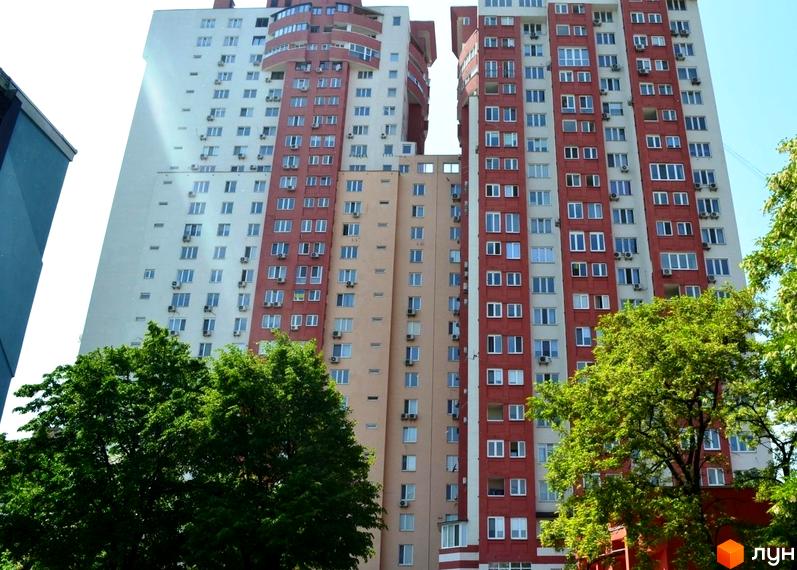 Багатоповерховий житловий комплекс на вул. Тичини, 18б з білими, червоними та бежевими фасадами. Навколо будівель ростуть зелені дерева.