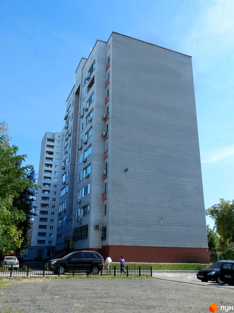 Зовнішній вигляд новобудови на вул. Вітряків (Маршала Судця), 7. Будинок цегляний, сірого кольору, з червоною цокольною частиною. Навколо будинку розташована прибудинкова територія з деревами та автомобілями.