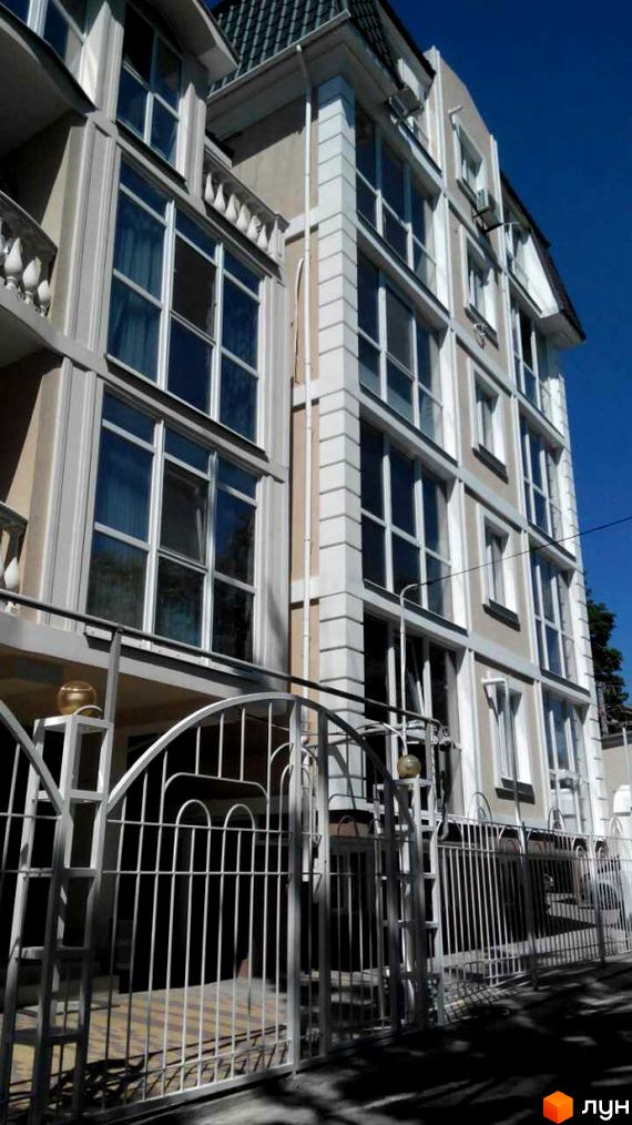 Зовнішній вигляд клубного будинку з великими вікнами та білими декоративними елементами. Перед будинком розташована кована огорожа та доглянута територія.