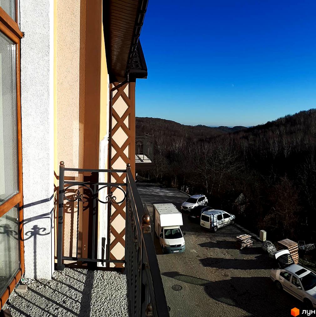 Фасад будинку світлого кольору з декоративними коричневими елементами та балконом з кованими перилами. На прибудинковій території видно парковку та лісисті пагорби.