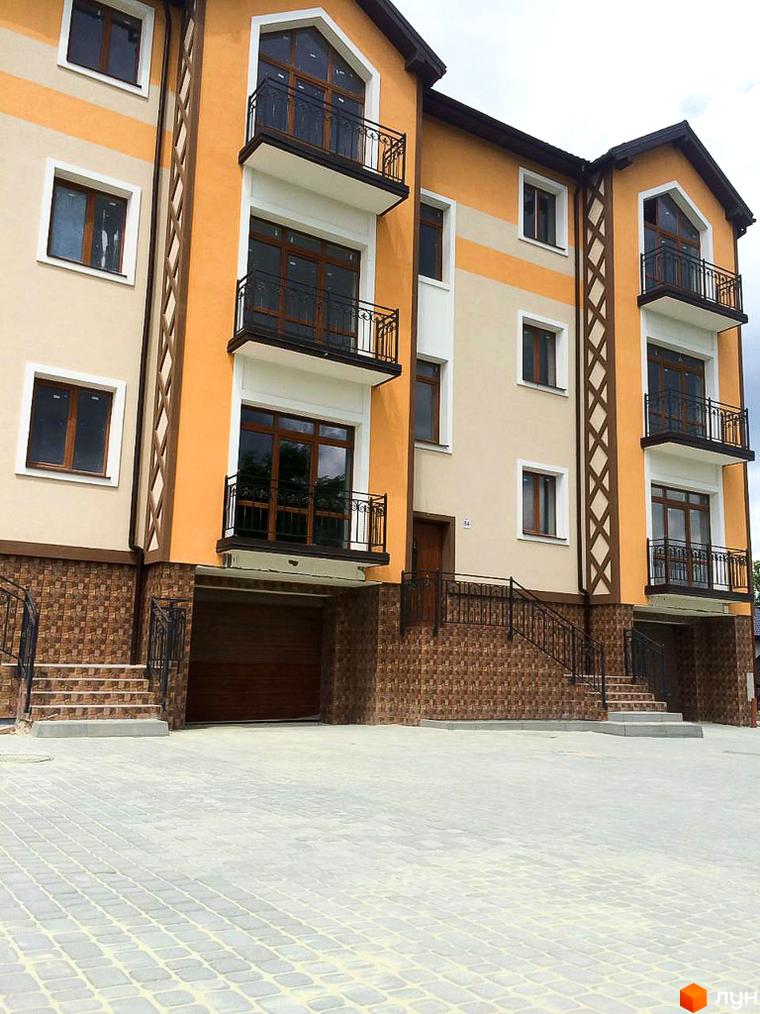Багатоквартирний будинок з помаранчево-бежевим фасадом, коричневими вікнами та балконами з металевими поручнями. Перед будинком вимощена світлою бруківкою територія та сходи до входу.