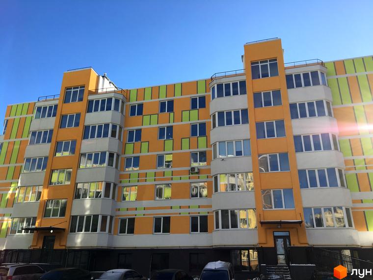 Житловий комплекс Позитив. Фасад будинку з яскравими смугами оранжевого, зеленого та білого кольорів. Поруч видно прибудинкову територію з автомобілями.