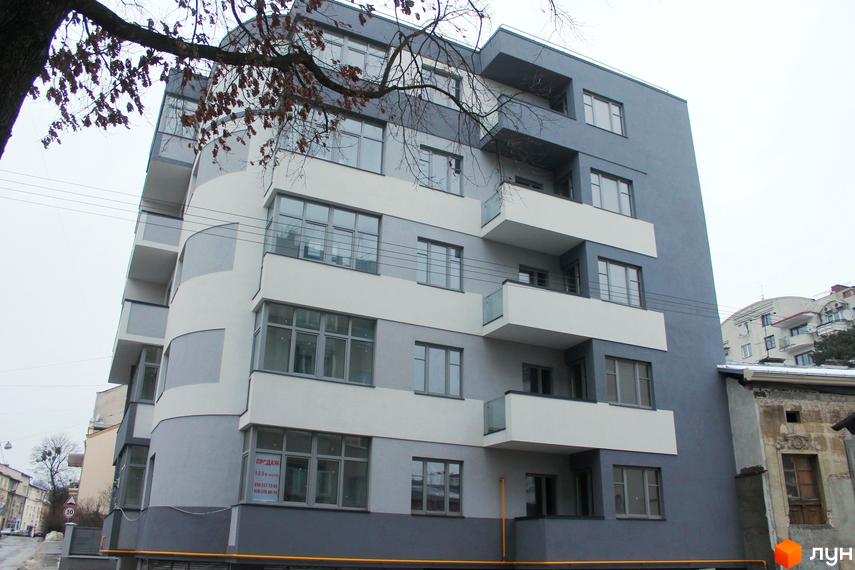 Багатоповерховий житловий будинок Клубний будинок Modern Lux з сіро-білим фасадом, вигнутими елементами та балконами. На передньому плані видно дерево без листя та міську вулицю.