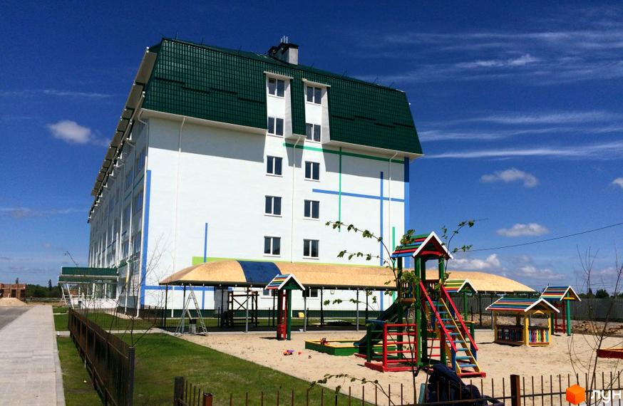 Новобудова ЖК Милі квартири з білим фасадом та зеленим дахом. Поруч дитячий майданчик з пісочницею та гірками.