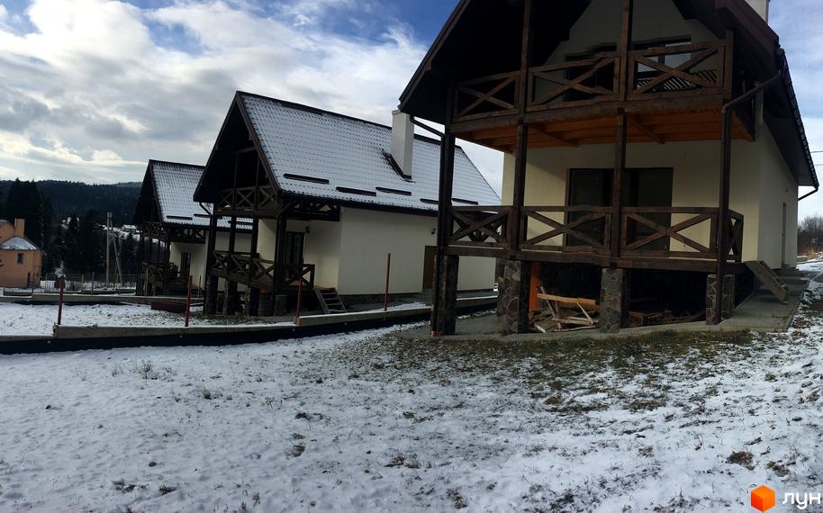 Зовнішній вигляд котеджів з дерев'яними балконами та дахами, вкритими снігом. Прибудинкова територія засніжена, з елементами ландшафтного дизайну.