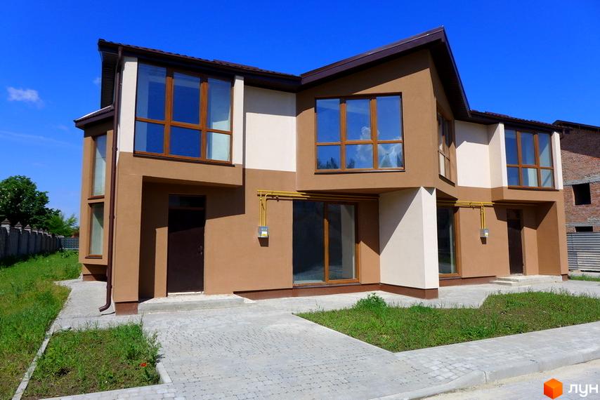 Двоповерховий таунхаус з великими вікнами та коричневою покрівлею. Перед будинком викладена бруківка та є газон.