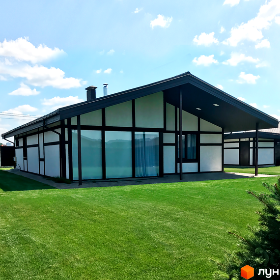 Білий будинок з темним дахом та вікнами. На передньому плані зелена трава, на задньому - блакитне небо з хмарами. Котеджне містечко Європейський квартал.