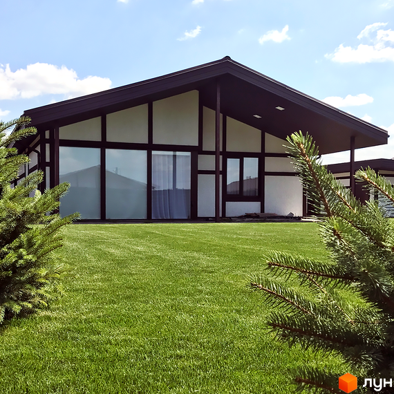 Будинок з великими вікнами та темним дахом в Котеджному містечку Європейський квартал. Зелена трава та кущі на прибудинковій території.