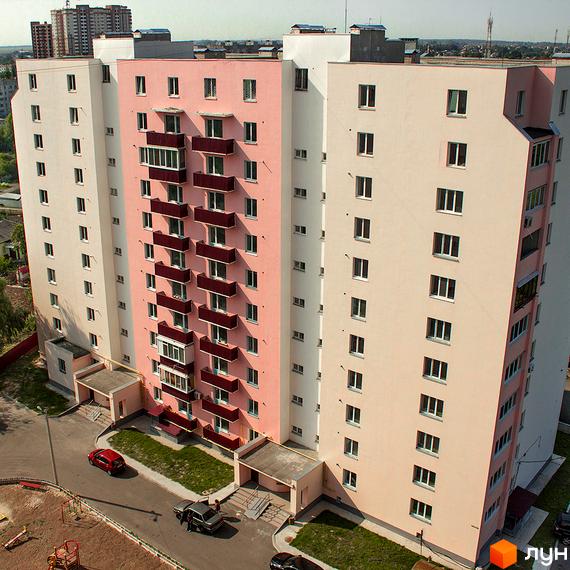 Зовнішній вигляд новобудови ЖК Нове Життя з рожевим та білим фасадами, балконами та вікнами. Прибудинкова територія з дорогою, зеленими насадженнями та дитячим майданчиком.