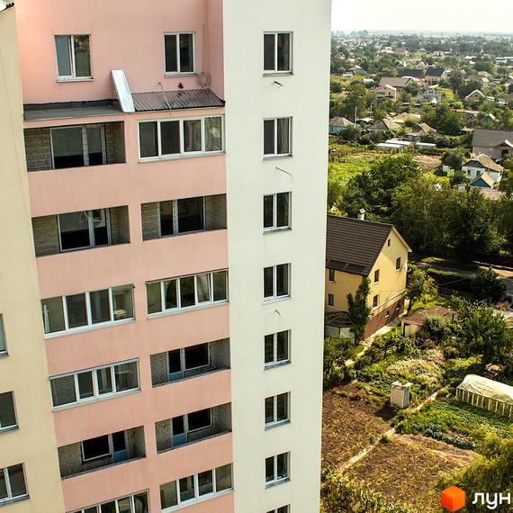 Зовнішній вигляд багатоповерхового будинку ЖК Нове Життя з рожевим та білим фасадами. Навколо будинку зелена прибудинкова територія з садами та сусідніми будинками.