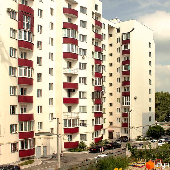 Житловий комплекс Лелека-1 з білими фасадами та червоними балконами. Прибудинкова територія з парковкою та зеленими насадженнями.