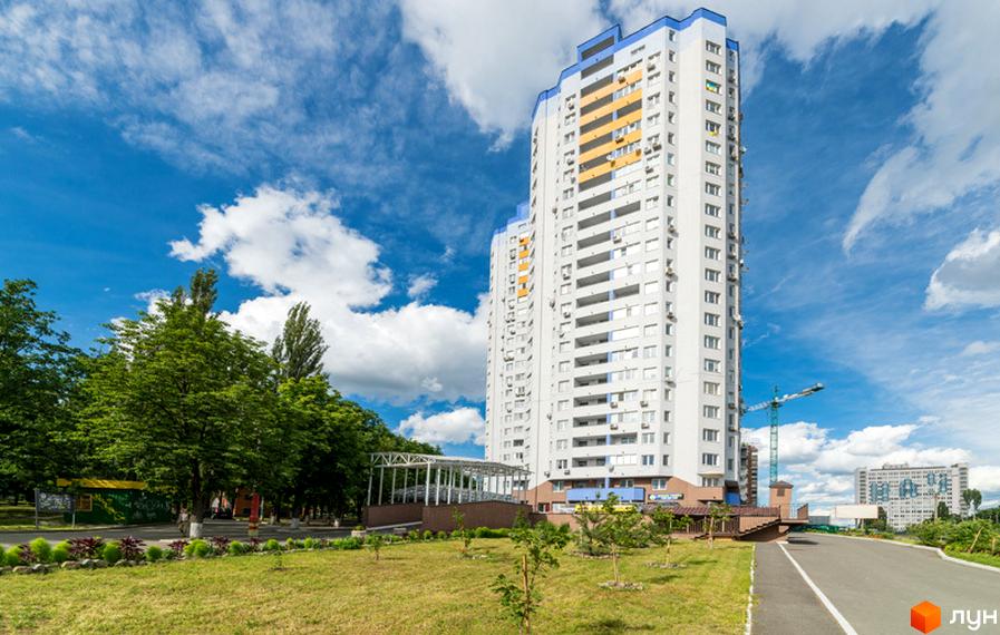 Житловий комплекс Авіатор: високий будинок білого кольору з синіми та жовтими елементами, оточений зеленими деревами та доріжкою.